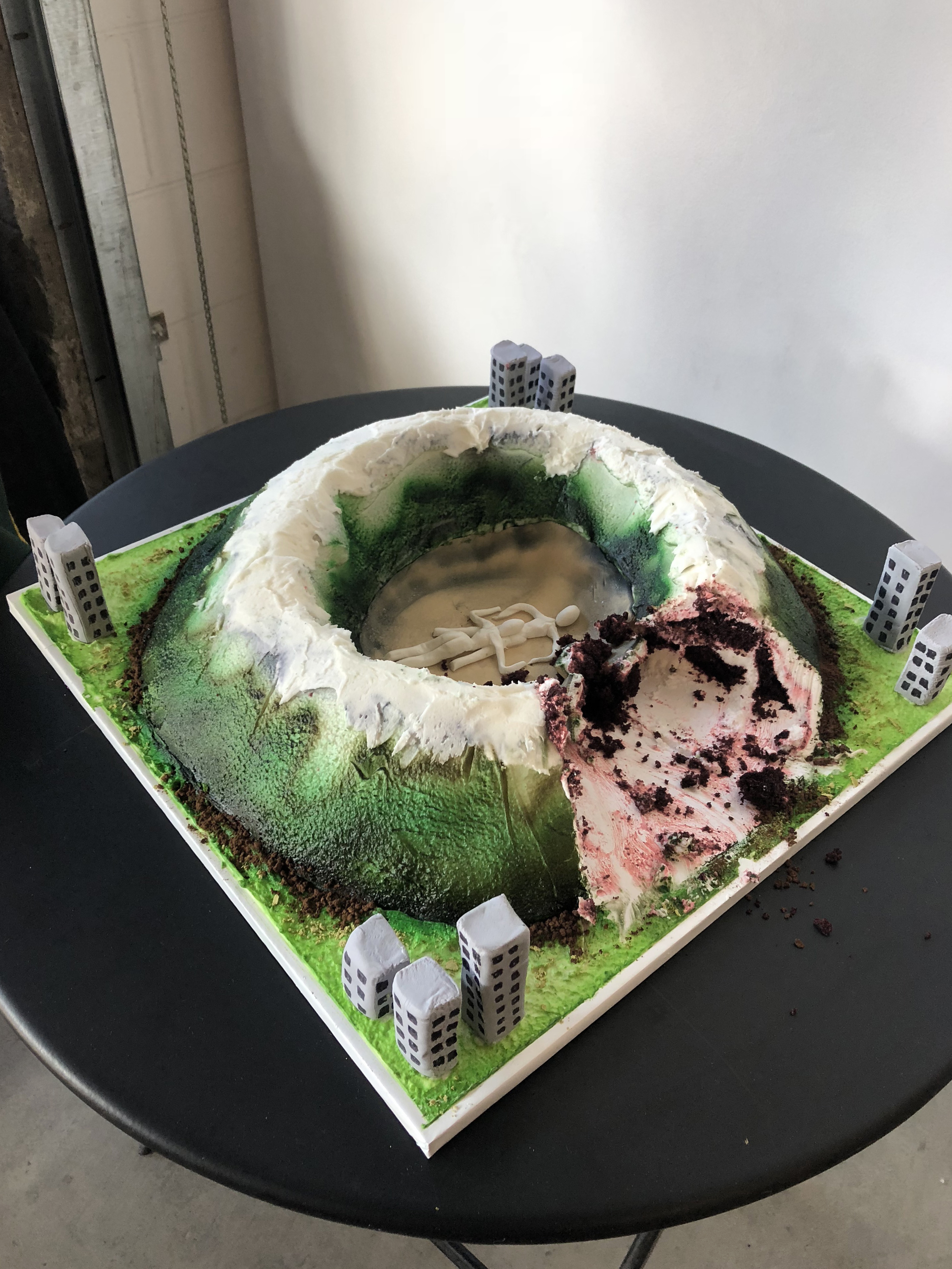 A cake shaped like a mountainous crater with two figures engaged in felatio in the center surrounded by buildings with a big slice cut out on a black table